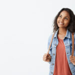 Cheerful gorgeous young Afro-American woman wearing jeans jacket and red t-shirt with dark hair smiling dreamily while thinking about something pleasant. Pretty girl dressed casually with excited joyful smile.