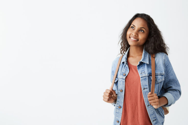 Cheerful gorgeous young Afro-American woman wearing jeans jacket and red t-shirt with dark hair smiling dreamily while thinking about something pleasant. Pretty girl dressed casually with excited joyful smile.