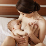 Young beautiful naked mom breastfeeding hugging her newborn baby smiling sitting on bed at home.