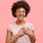 Photo of friendly positive female smiles sincerely, has Afro hair, keeps both palms on heart, dressed in casual t shirt, isolated over pink wall, expresses good feelings and attitude to close person