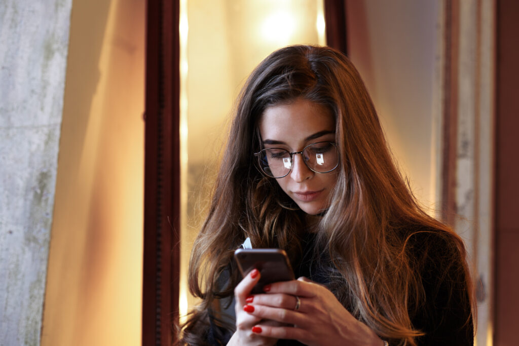 Headshot of beautiful young European woman wearing eyeglasses and black dress using mobile phone indoors, messaging friends online or browsing her newsfeed via scoial networks. Staying connected