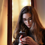 Headshot of beautiful young European woman wearing eyeglasses and black dress using mobile phone indoors, messaging friends online or browsing her newsfeed via scoial networks. Staying connected