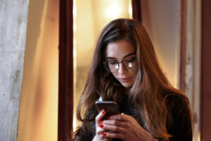 Headshot of beautiful young European woman wearing eyeglasses and black dress using mobile phone indoors, messaging friends online or browsing her newsfeed via scoial networks. Staying connected